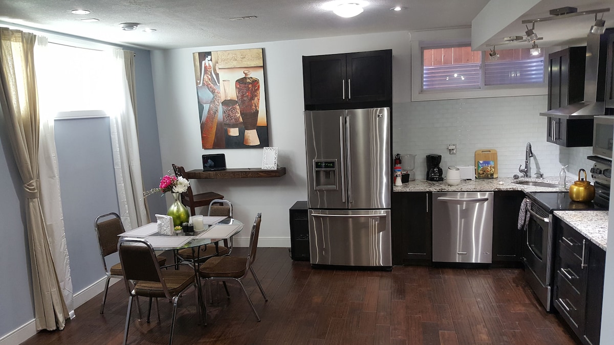 The image displays a modern kitchen and dining area, featuring stainless steel appliances, including a refrigerator and a stove. A small dining table with four chairs is set beside a window adorned with light curtains. A painting and decorative items add visual interest to the space.