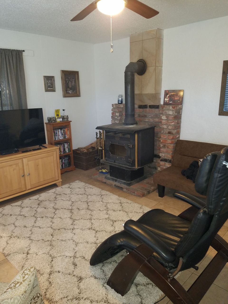 A cozy living area is presented with a plush area rug and a reclined black chair. A wooden television cabinet stands opposite a vintage wood-burning stove, which is set against a brick backdrop. Soft lighting is provided by a ceiling fan above.