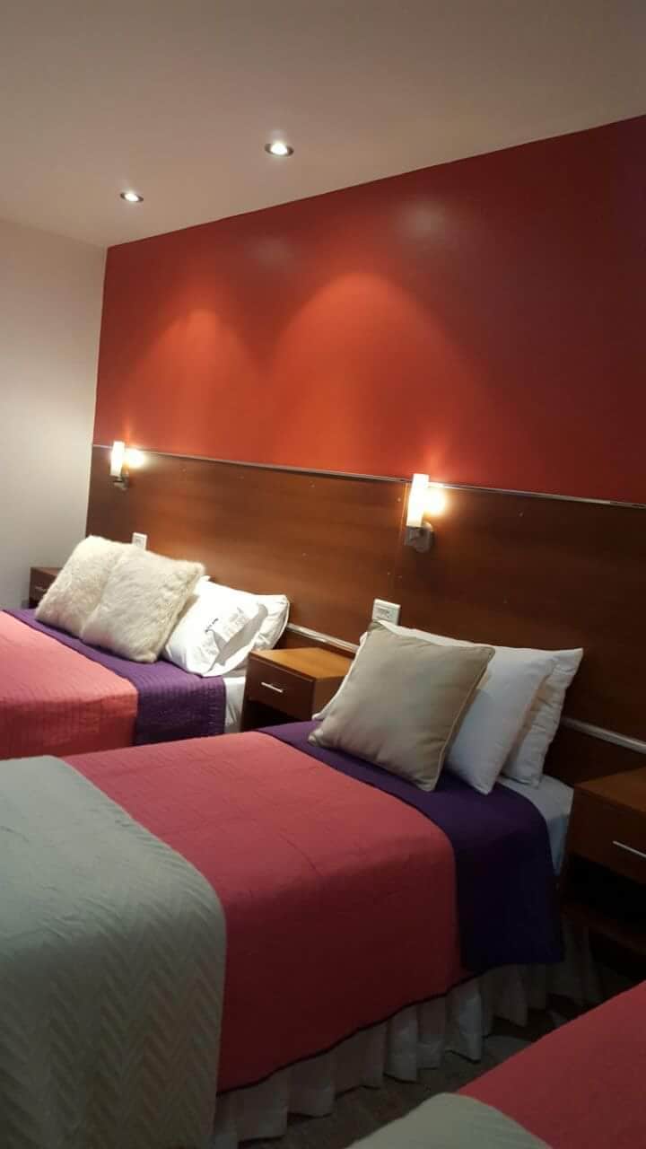 A sleeping area is shown with three beds, each topped with distinct blankets in pink, purple, and light green. Soft pillows are arranged at the head of each bed, while wall-mounted lights provide ambient illumination. The walls are painted in a warm red tone, enhancing the overall warmth of the space.