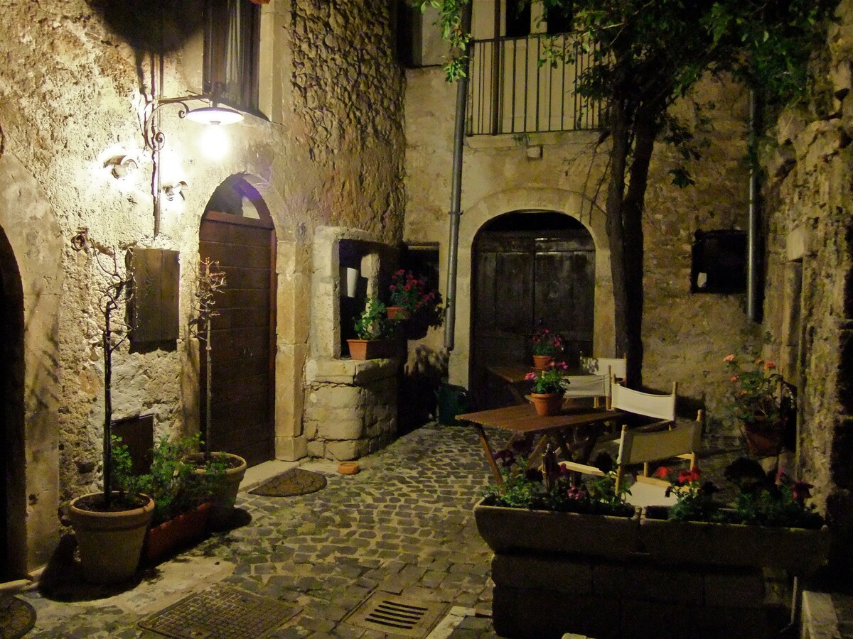 A quaint courtyard is illuminated by soft lighting, featuring stone walls and a cobblestone path. Potted plants add vibrant greenery near a small wooden table and chairs, creating a cozy outdoor seating area. Arched doorways and rustic doors enhance the historical charm of the setting.