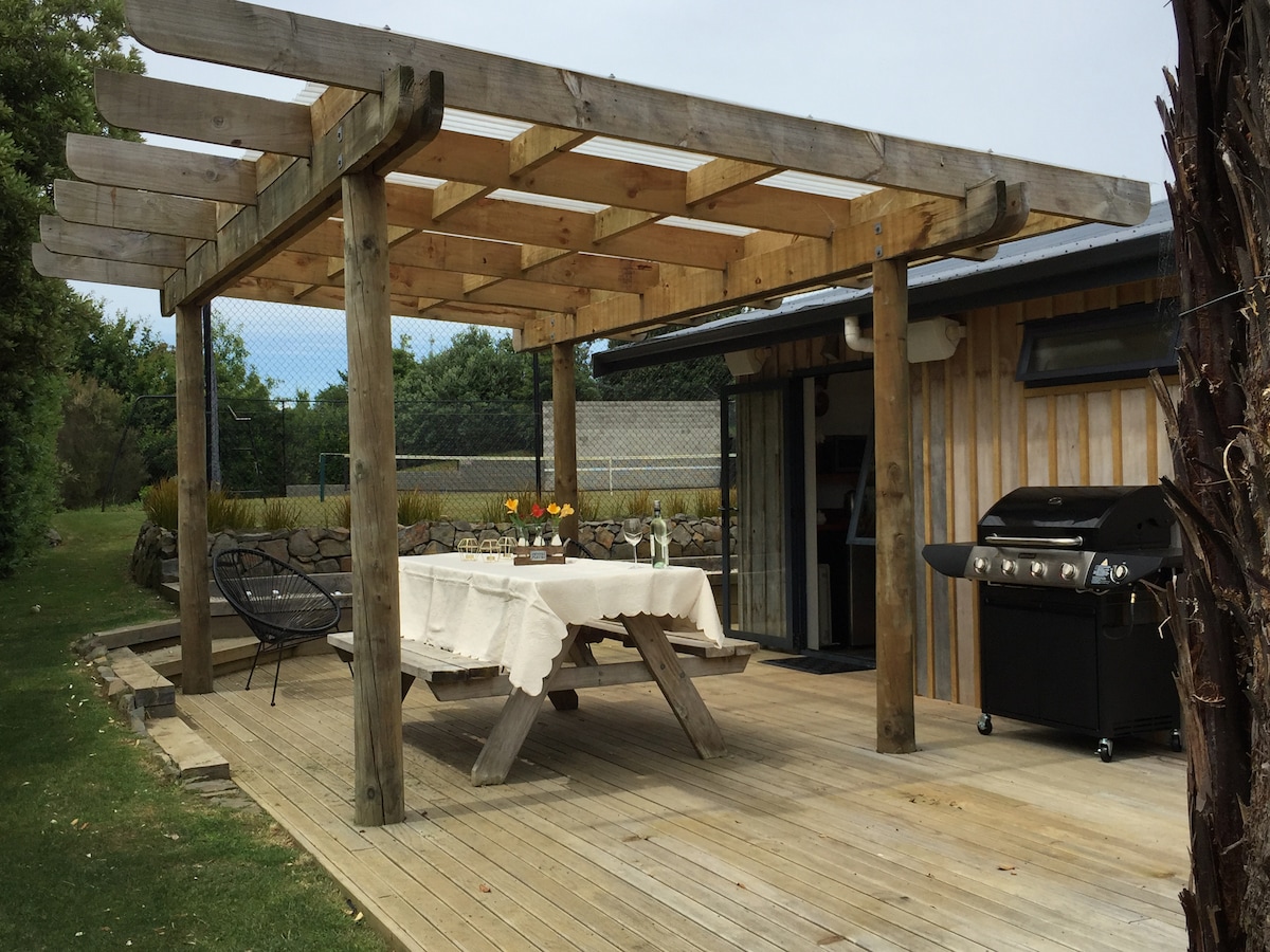 An outdoor deck area is shown, featuring a large picnic table with a light-colored tablecloth and a barbecue grill. Surrounding seating is included, and a trellis partially provides shade overhead. The space is adjacent to a grass tennis court, enhancing the recreational atmosphere.