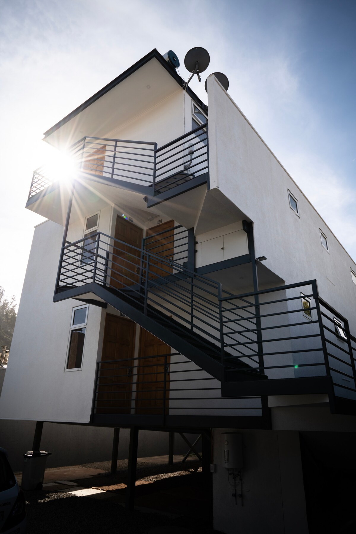 The modern building features a minimalist design with a two-story structure. A staircase with metal railings leads to an upper level. Large windows allow natural light to fill the space, while satellite dishes are mounted on the roof, capturing a clear blue sky.