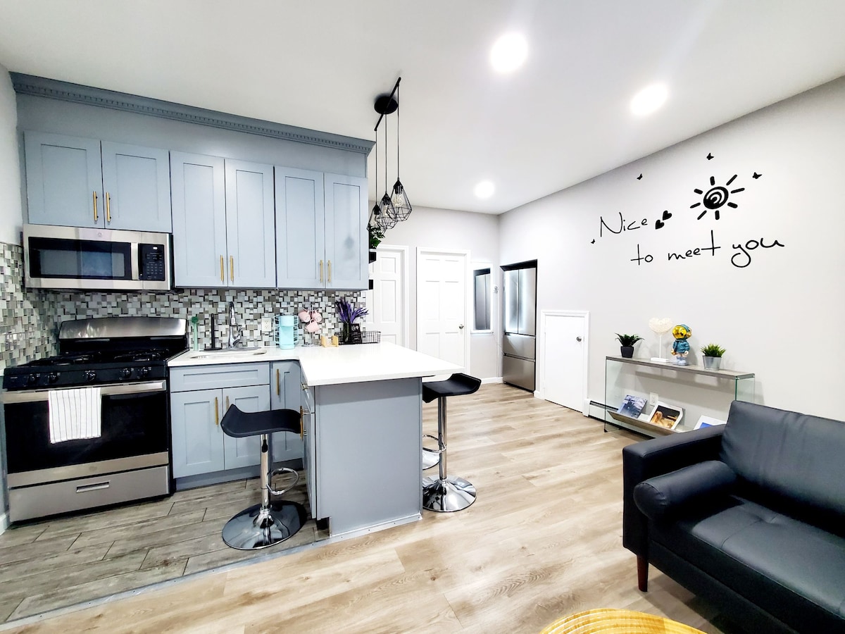 The modern kitchen features light blue cabinets and a center island with seating for two. Stainless steel appliances are present, along with a decorative backsplash. A welcoming message is displayed on the wall, while a cozy seating area is positioned nearby.