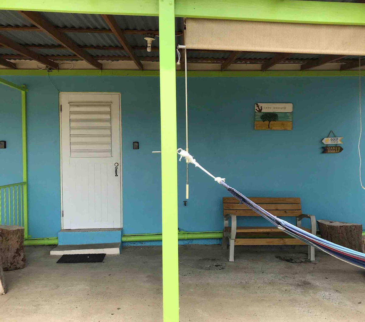 A vibrant blue exterior is showcased, featuring a wooden door with a simple lock. A welcoming patio is highlighted with a hammock hanging between two support posts and a bench positioned against the wall under a roof. Natural wooden elements are present on the ground.