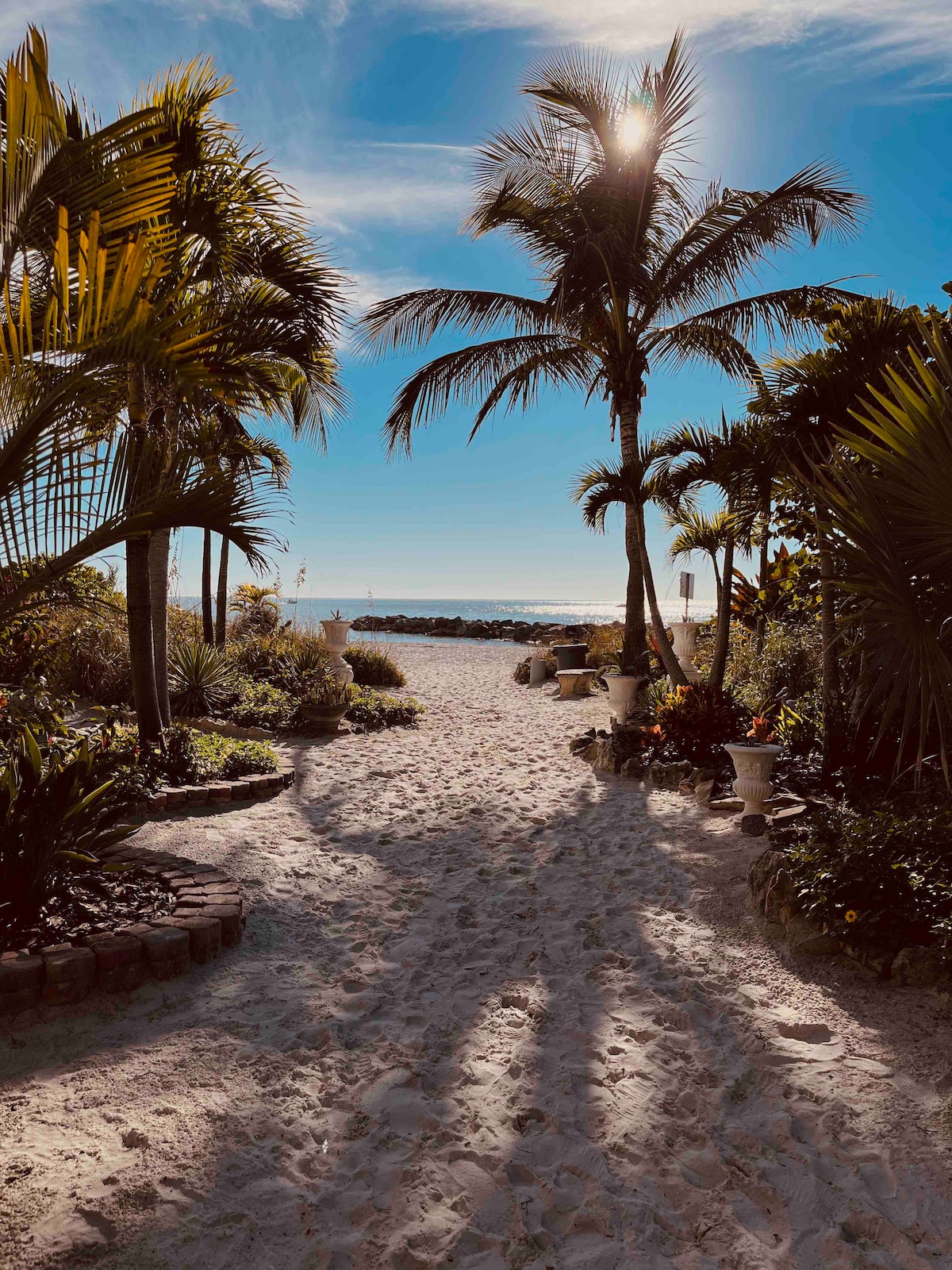 A sandy path leads through lush tropical plants, creating a serene entrance to the beach. Tall palm trees provide shade as sunlight filters through, casting gentle shadows on the ground. The distant coastline is visible, hinting at a peaceful beach experience.