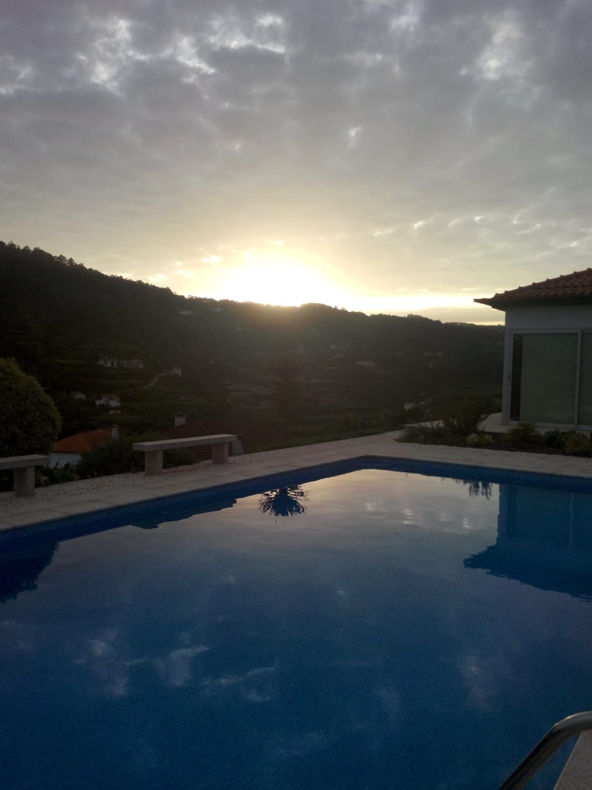 A clear pool reflects the vibrant colors of the sky at sunrise, surrounded by lush hills. The scene is framed by a nearby house, with greenery enhancing the natural setting.
