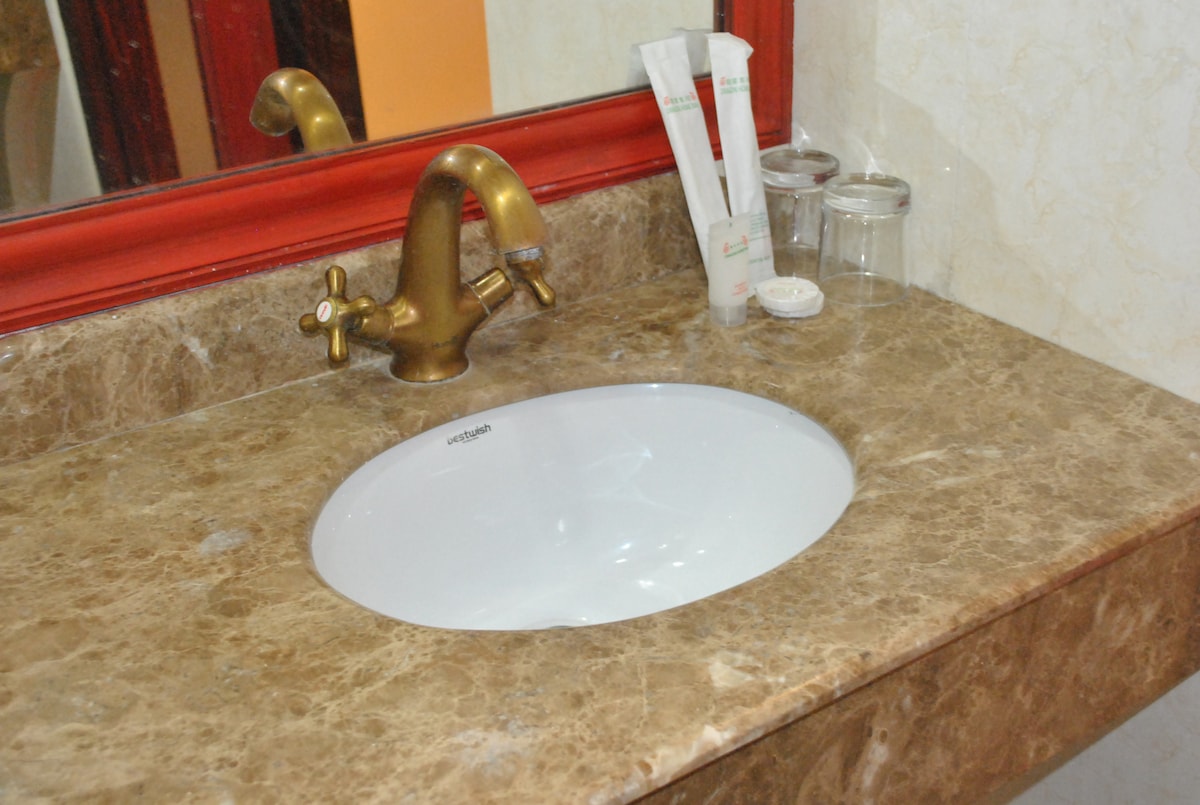 A bathroom sink is positioned on a marble countertop, featuring a curved white basin and a brushed gold faucet. Two glass containers and a set of toiletries are arranged neatly beside the sink. A decorative red mirror frame reflects the space.