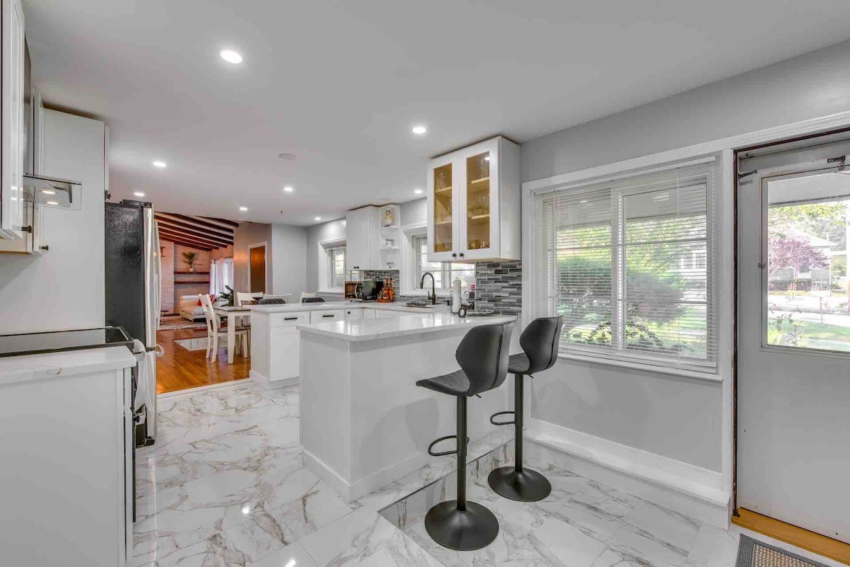 The modern kitchen features a central island with two bar stools positioned nearby. Light-coloured cabinetry complements the marble-patterned flooring. Large windows allow natural light to fill the space while offering views of the outdoor yard. A doorway leads to a welcoming dining area.