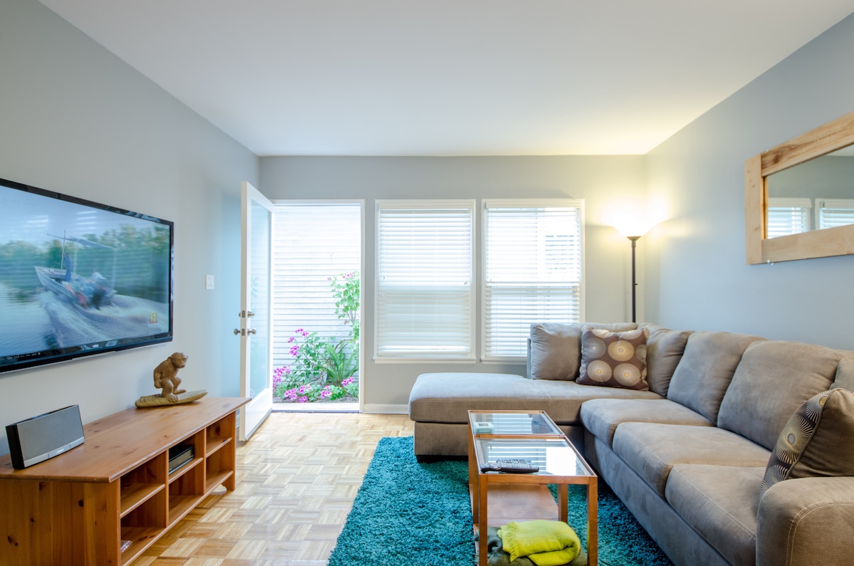 A comfortable living area is equipped with a large sectional sofa, a coffee table, and a television mounted on a wooden media unit. Natural light filters through the multiple windows, highlighting the parquet flooring and a vibrant area rug beneath the furniture.