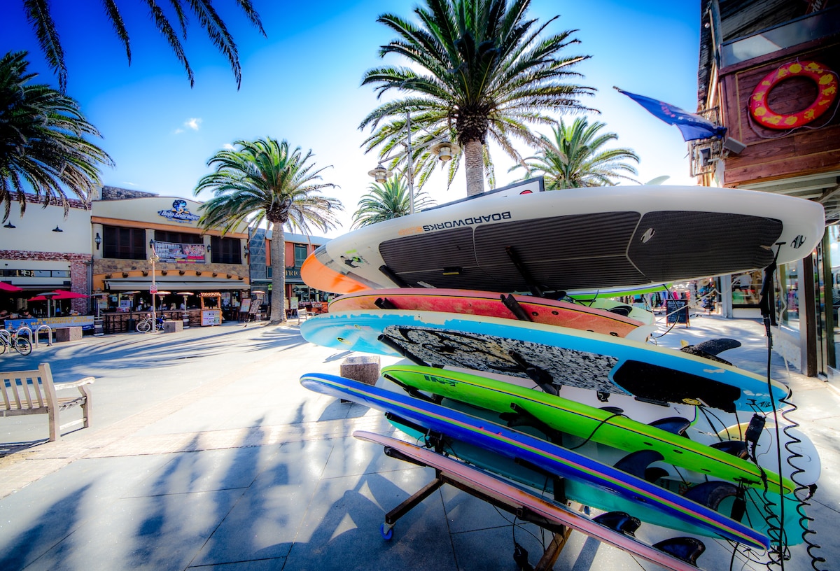 Several colorful surfboards are stacked vertically against an outdoor vendor stall, surrounded by palm trees. The vibrant colors of the surfboards create a lively atmosphere, while the pedestrian walkway leads toward shops and restaurants in the background.