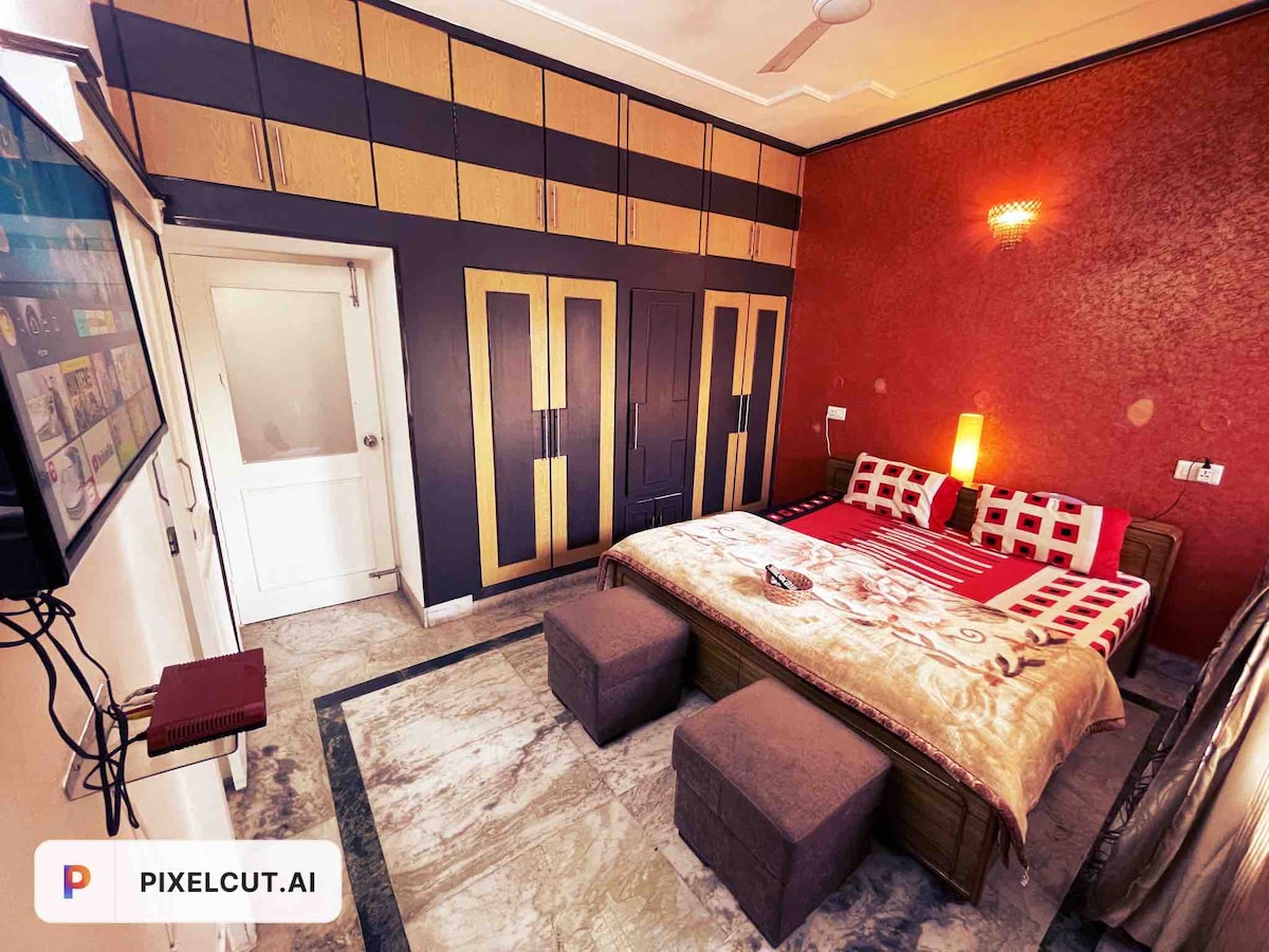 The bedroom features a spacious bed with a patterned coverlet, complemented by two upholstered stools positioned at the foot. The walls showcase a bold red hue, along with stylish cabinets that provide ample storage. A television is mounted on the wall, enhancing entertainment options.