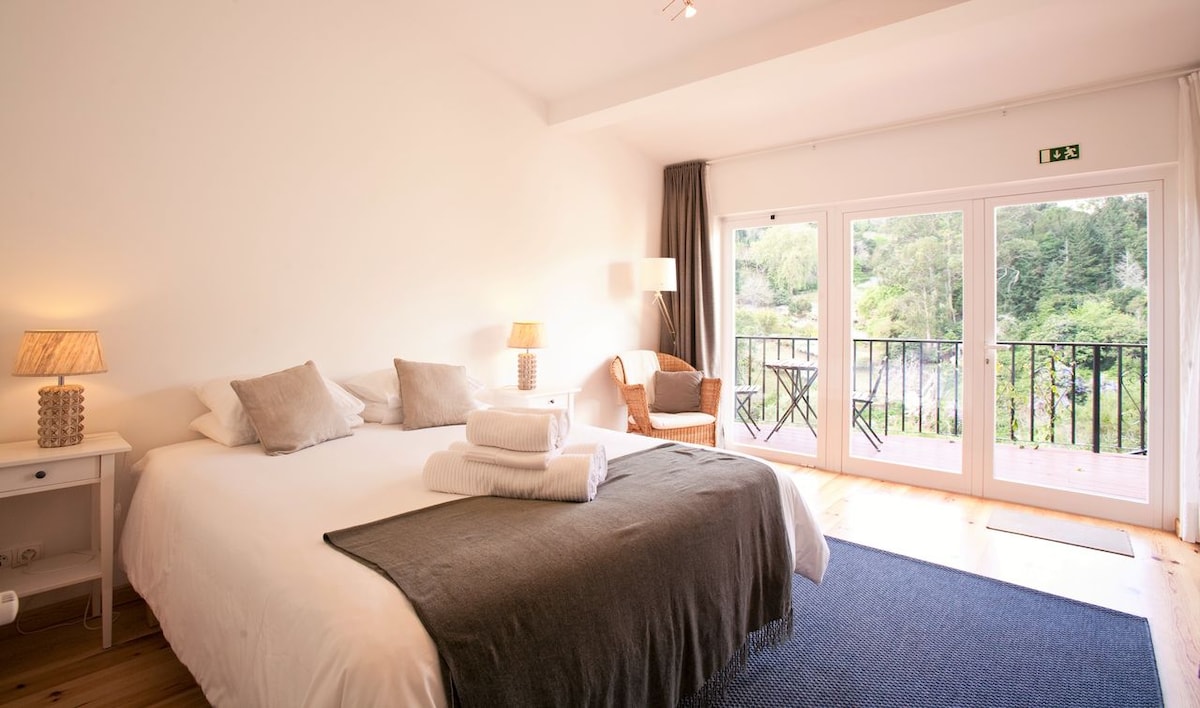 A spacious bedroom features a large bed draped with soft linens, complemented by decorative pillows. Two bedside lamps provide gentle illumination. A seating area with a cozy chair and a small table is visible, along with a balcony offering views of greenery through glass doors.