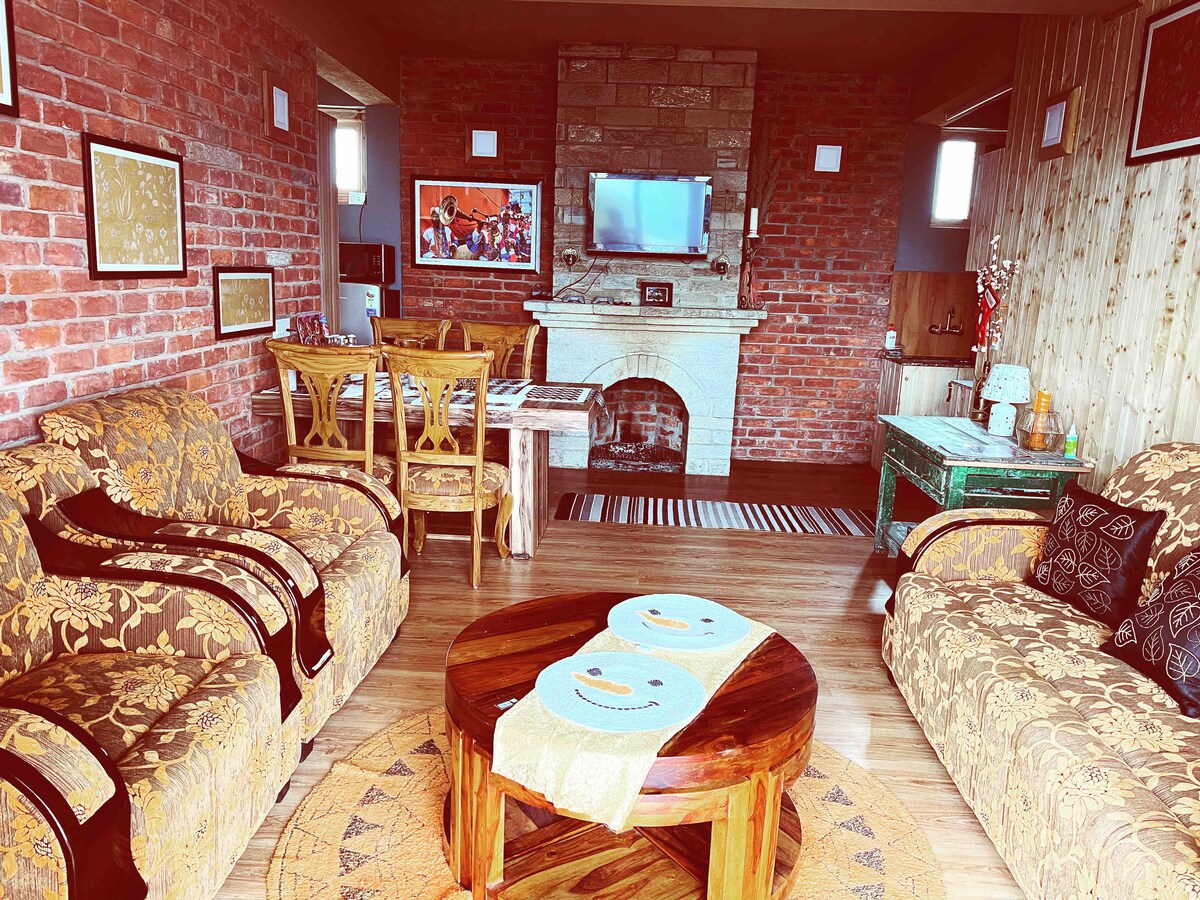 A living area is presented with a combination of plush seating arrangements, including a patterned sofa set and wooden chairs. A coffee table is centered on a decorative rug, while a stone fireplace adds warmth to the space. Natural light filters in through windows adorned with light curtains.