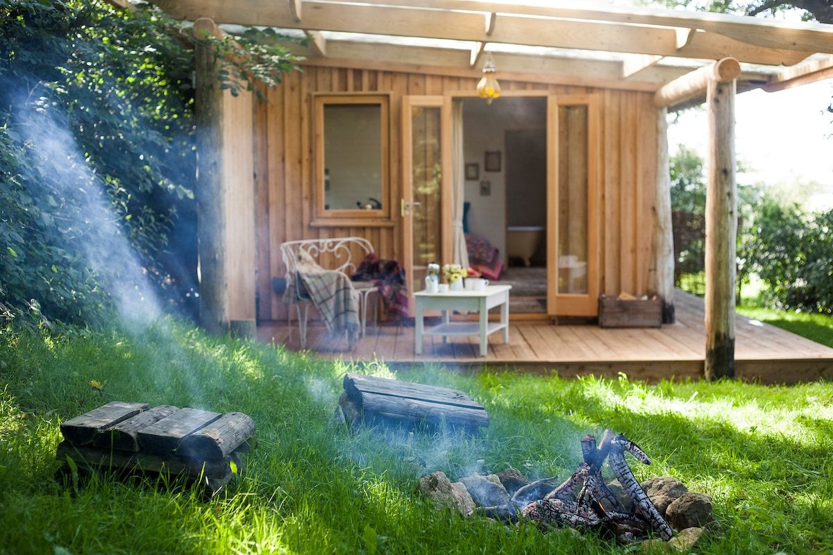 A wooden cabin is visible with large glass doors opening onto a deck. A small table and chairs are positioned nearby, offering a place to relax. In the foreground, a fire pit with gently smoking logs adds to the outdoor ambiance, surrounded by lush green grass.