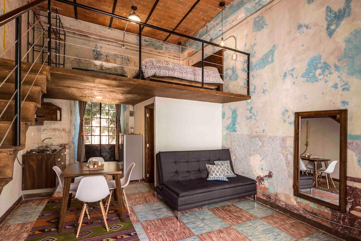 A spacious studio apartment features a wooden loft with a bed above. The main area includes a comfortable sofa bed and a dining table with modern chairs. The walls display a charming mix of distressed paint, while a large mirror enhances the sense of space.