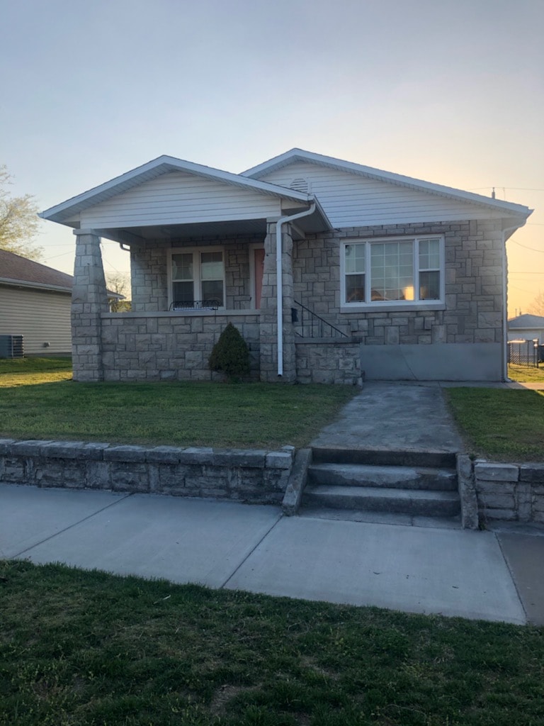 The exterior of the cottage is shown, featuring stone facade and a sloped roof. A small porch is visible, with steps leading up to the entrance. A well-kept lawn surrounds the house, and a concrete walkway provides access from the street.