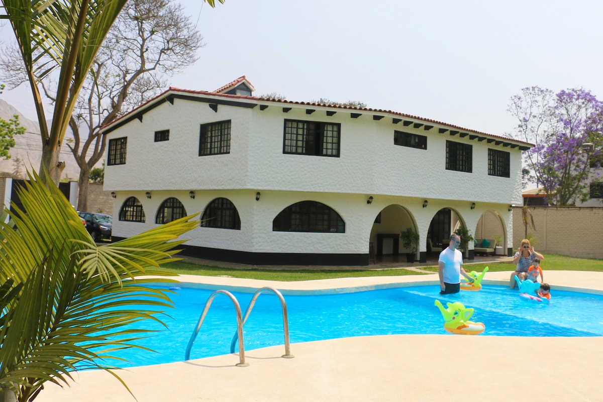 A spacious two-story building with a white exterior features arch-shaped windows. In the foreground, a pool with clear blue water is accompanied by guests enjoying floatation toys, surrounded by palm trees and a landscaped area.