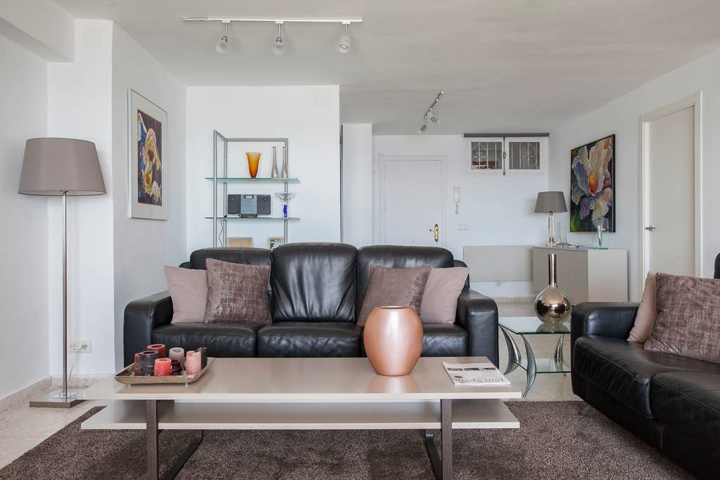 A spacious living room features a pair of black leather sofas adorned with neutral-colored cushions. A modern coffee table sits in the center, complemented by a decorative bowl. The light, airy atmosphere is enhanced by artwork displayed on the walls and a glass shelving unit.