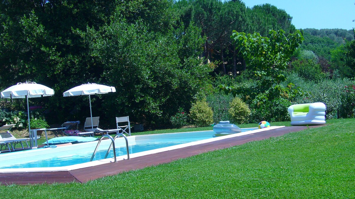 A private swimming pool is surrounded by a spacious lawn, featuring lounge chairs and shaded umbrellas. Green trees and shrubs provide a serene backdrop, while colorful pool floats rest on the water's surface. The area offers an inviting space for relaxation and leisure.
