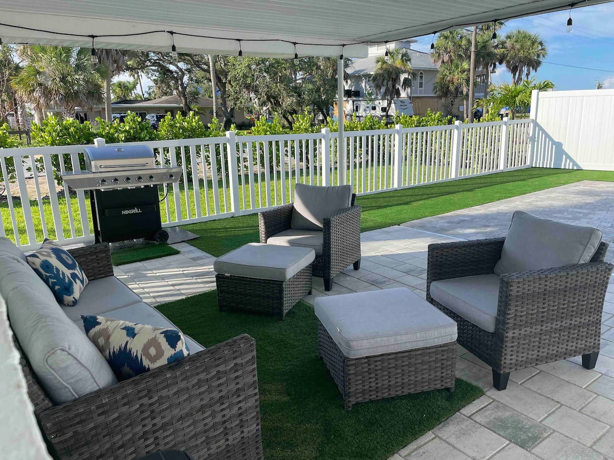 An outdoor lanai features a comfortable seating arrangement with a sectional sofa, chairs, and ottoman, all on a grassy surface. A grill is positioned nearby, and the area is surrounded by a white fence, with palm trees visible in the background.