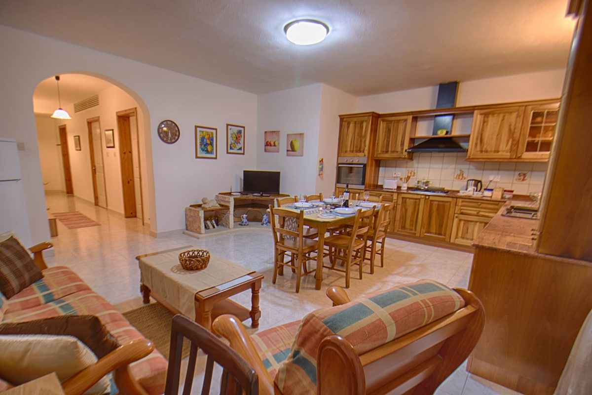 The open living area features a wooden dining table seating six, surrounded by a comfortable sofa and chairs. A television is positioned on a nearby stand, while the kitchen showcases wooden cabinets and a countertop equipped for meal preparation. Art pieces adorn the walls.
