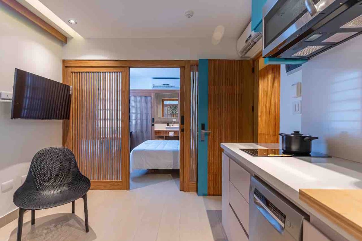 The interior space features a modern kitchen with stainless steel appliances and a sleek countertop. A black chair is placed adjacent to a wall-mounted television. Wooden sliding doors separate the kitchen area from a sleeping space, which is visible in the background.