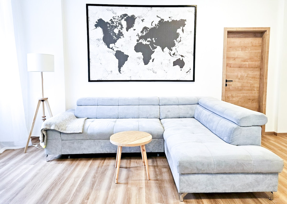 A light gray sectional sofa, positioned in a spacious living area, complements a world map artwork displayed above. A wooden round coffee table is placed in front of the sofa, enhancing the room's relaxed atmosphere. Soft natural light filters through nearby sheer curtains.