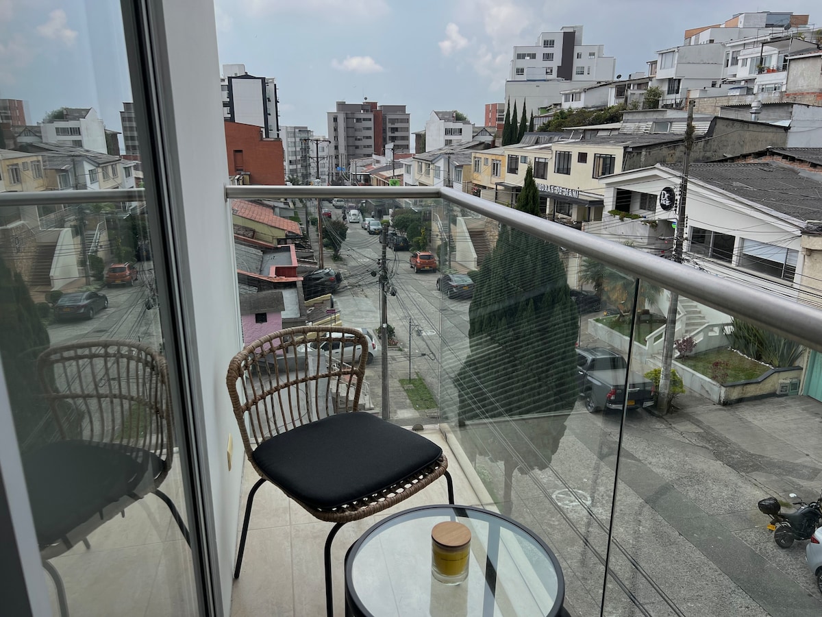 A balcony is seen featuring a round glass table beside a black chair. The view showcases a mix of buildings and greenery, with streets and parked cars visible in the distance. Natural light illuminates the space.