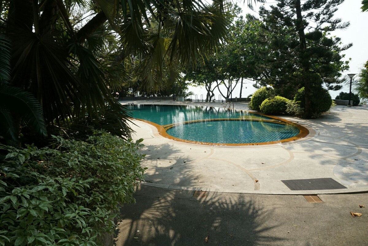 A tranquil swimming pool is framed by lush tropical foliage, including palm and shrubbery. The pool features a circular shape with a tiled surface reflecting the surrounding greenery, while a concrete area provides space for relaxation. A view of trees and water can be seen in the background.