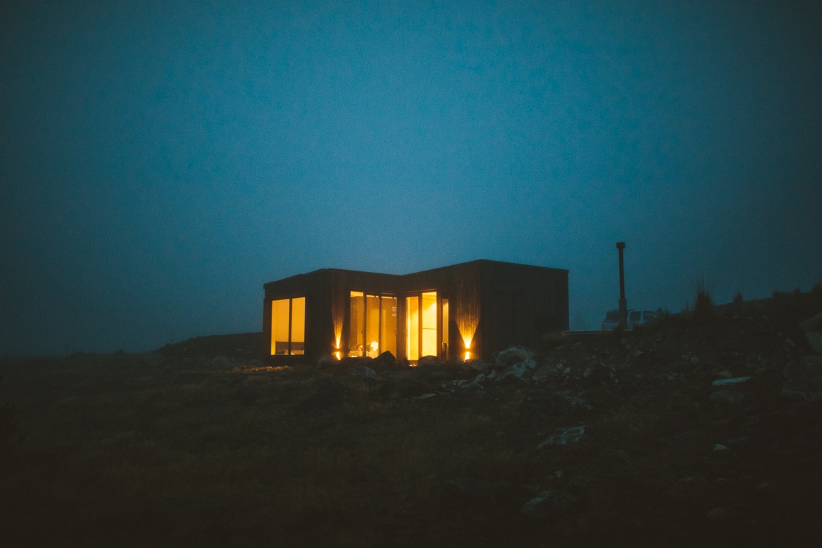 The cabin stands surrounded by nature during twilight, with warm light emanating from its large windows. A chimney is visible against the misty backdrop, contributing to the inviting appearance of the modern structure.