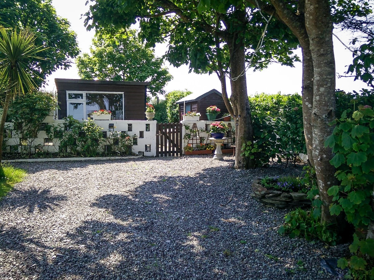 A gravel pathway leads to a wooden cabin surrounded by lush greenery, including decorative plants and trees. A simple white fence frames the entrance, adorned with flower pots. The serene outdoor area offers a welcoming first impression of the cozy cabin.