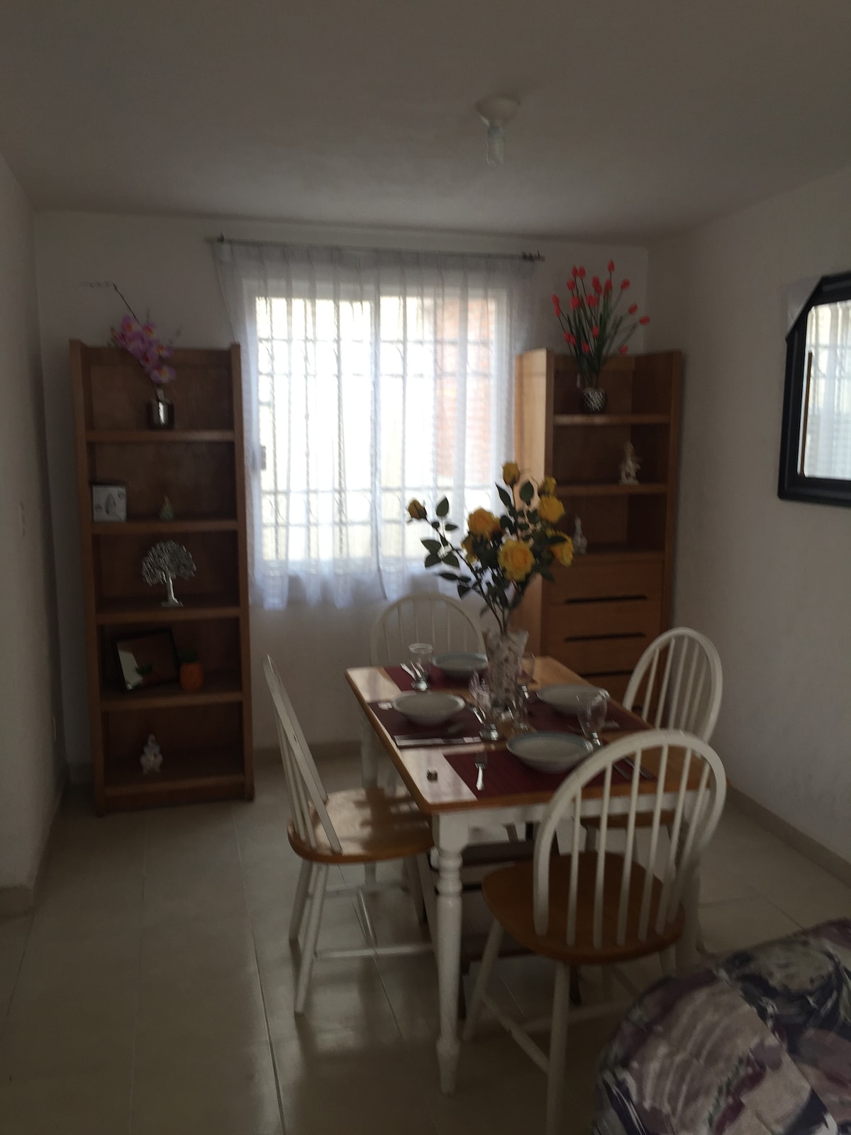 A dining area features a wooden table set for four, with white chairs and tasteful centerpieces. Natural light enters through a large window adorned with sheer curtains. Two wooden shelving units display decorative items and plants, enhancing the simplicity of the space.