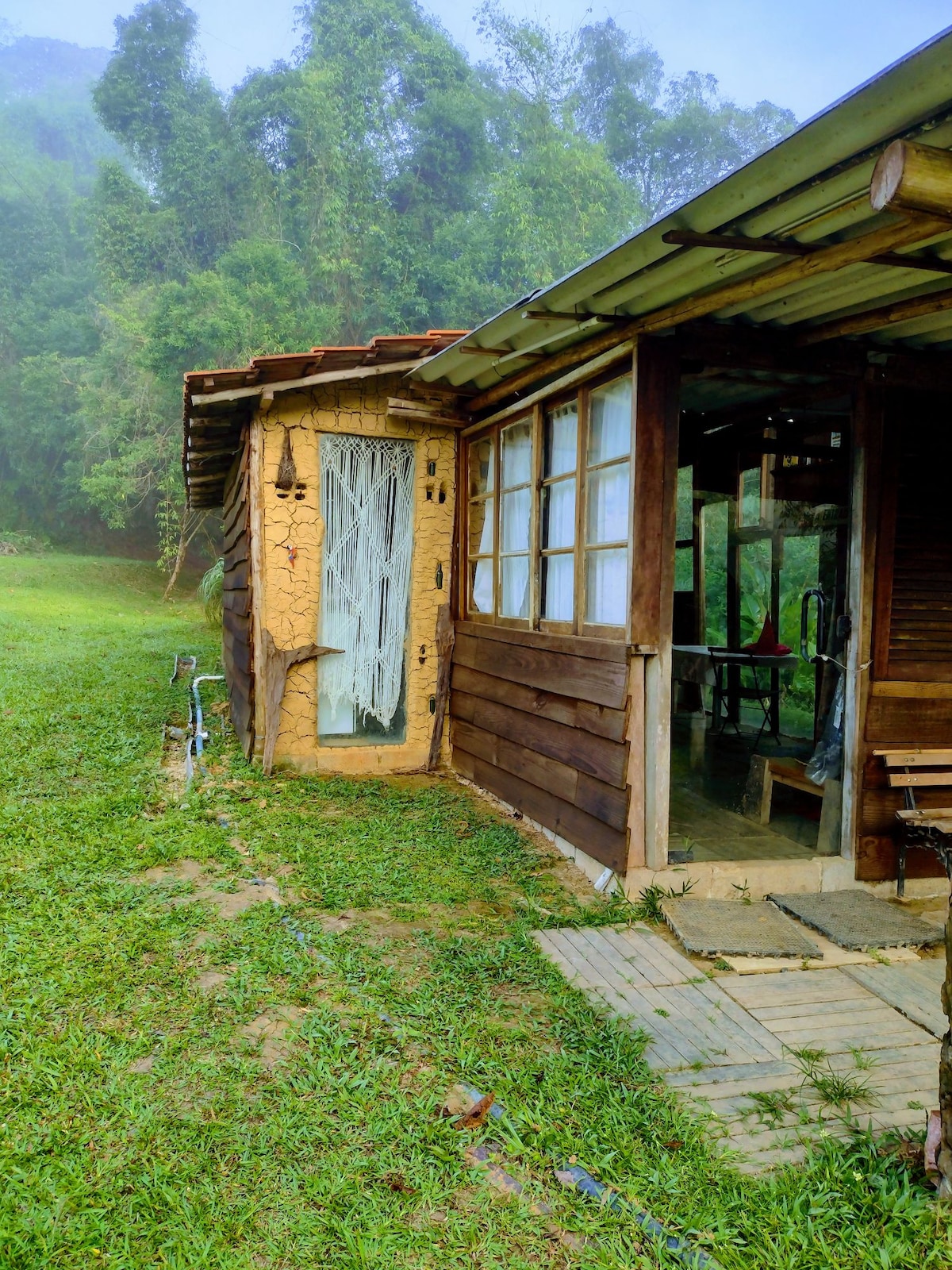 A rustic structure features a wooden exterior with a clay wall. Large windows provide views of the surrounding greenery, while a small porch area with wooden seating complements the natural landscape. The entrance showcases a decorative door and is framed by lush grass.