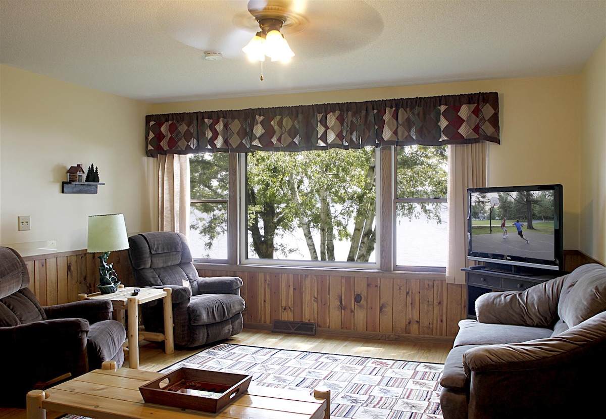 A living area features large windows offering a view of the lake. Comfortable seating is arranged around a wooden coffee table, with a television positioned nearby. Soft lighting is provided by a ceiling fan and a side lamp, enhancing the inviting space.