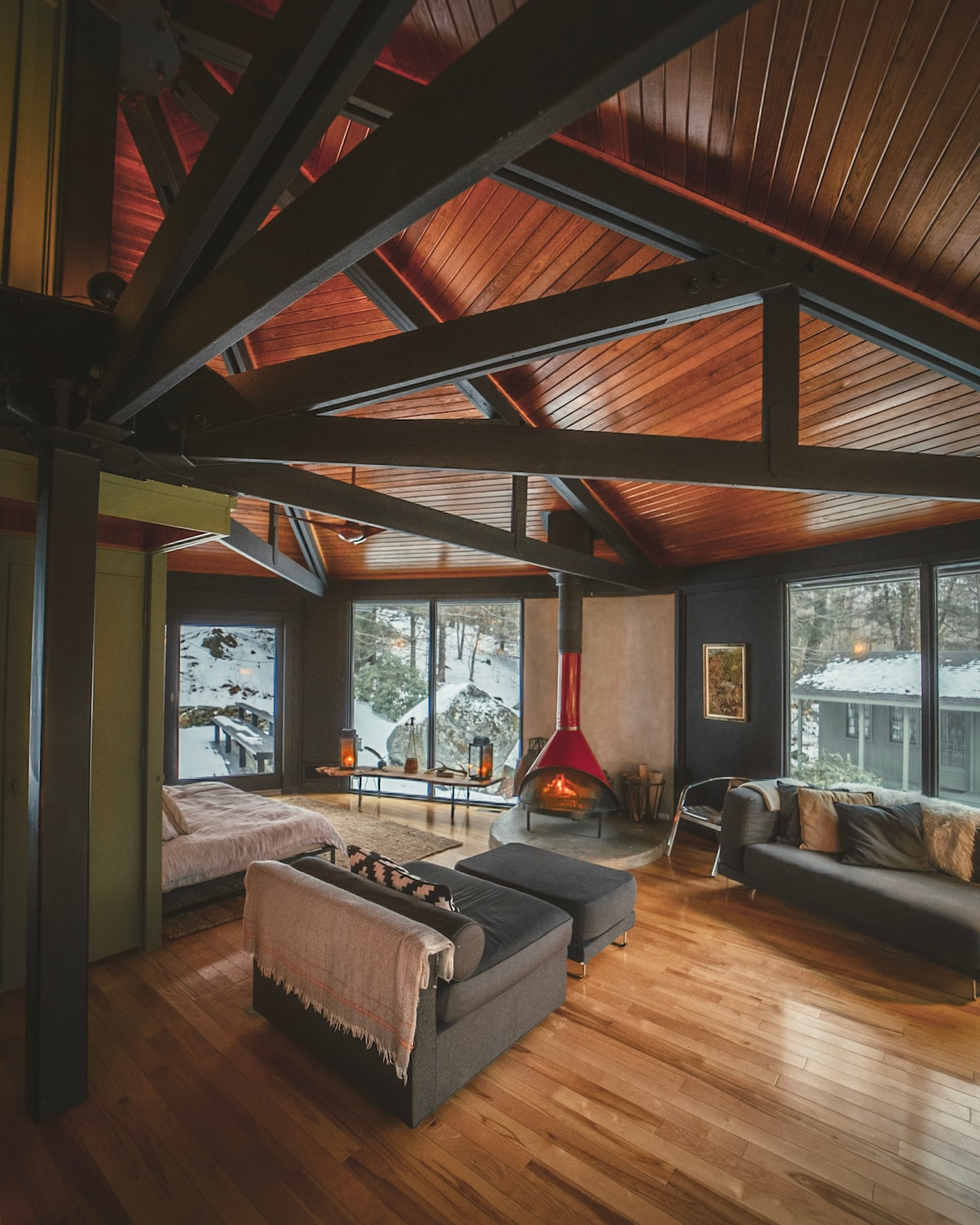 An inviting living area features vaulted wooden ceilings and large windows showcasing views of the surrounding nature. A modern wood-burning fireplace is prominently positioned, complemented by comfortable seating. Natural wood flooring adds warmth to the space, creating a cozy and open atmosphere.