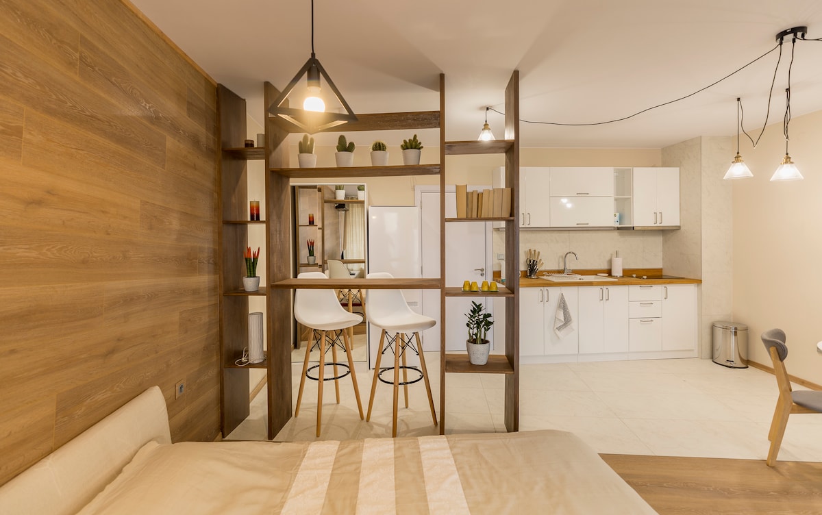A well-designed studio layout features a spacious bed in the foreground, with a wooden partition leading to a modern kitchen area. Two bar stools are positioned at a counter, enhancing the open-plan feel, while soft lighting accents the warm tones throughout the space.