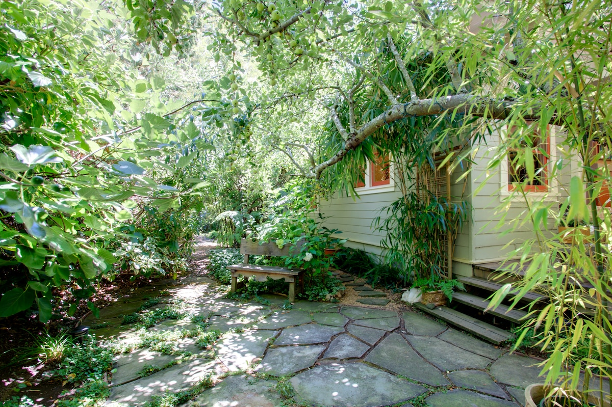 A tranquil garden surrounds the cottage, featuring natural stone pathways and lush green foliage. Overhead branches create a serene canopy, while a wooden bench offers a resting spot amidst the vibrant plants. The entrance to the cottage is framed by bamboo and flowering shrubs.