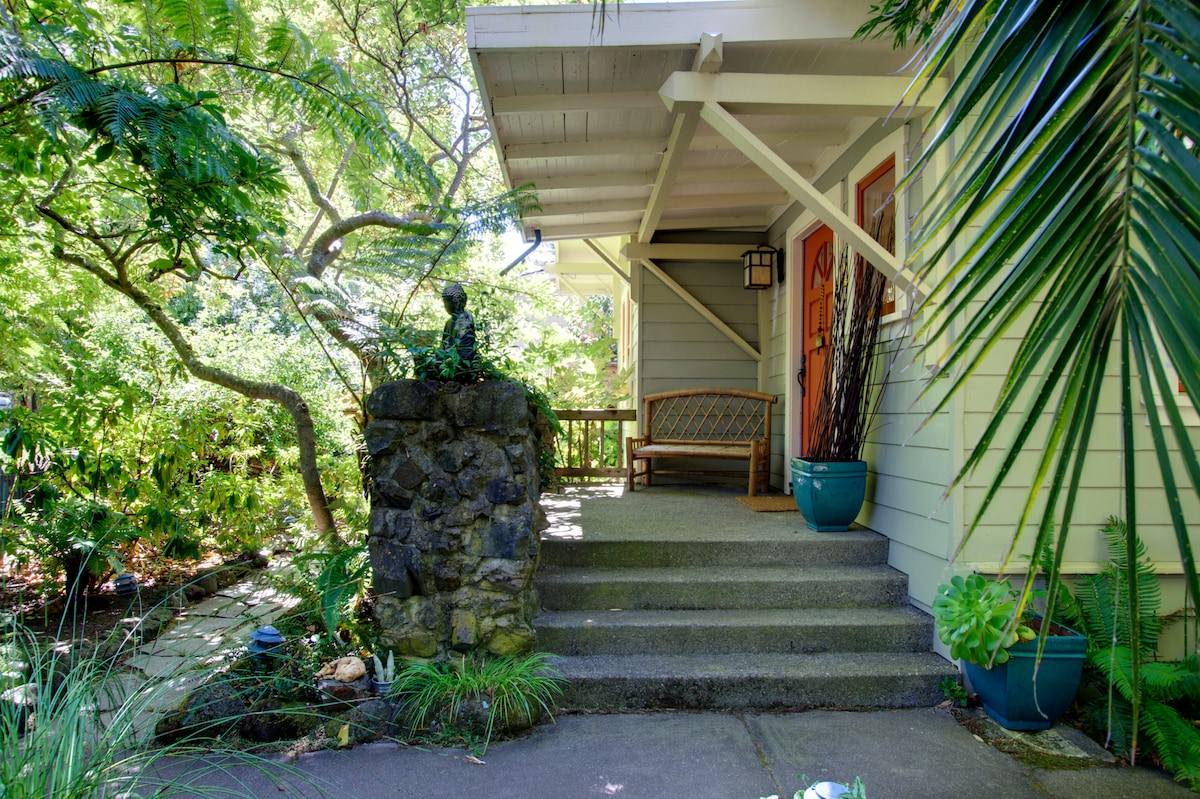 A welcoming entrance features a stone pathway leading to the front door. Lush greenery surrounds the area, with potted plants placed beside the steps. A wooden bench provides a cozy seating option, complemented by a decorative stone statue.