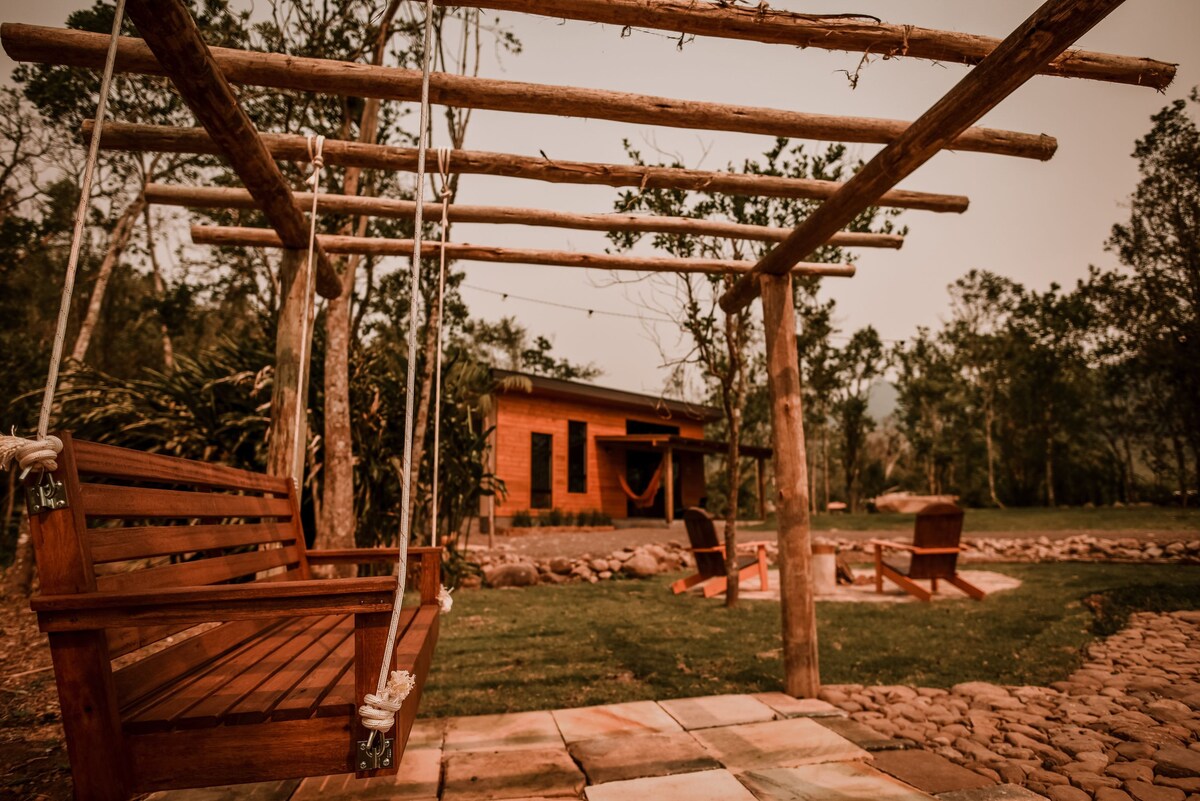 A wooden swing is suspended from a rustic frame in a green outdoor space. In the background, a cabin is visible surrounded by trees. A circular seating area with stone paving can be seen nearby, creating a serene environment.