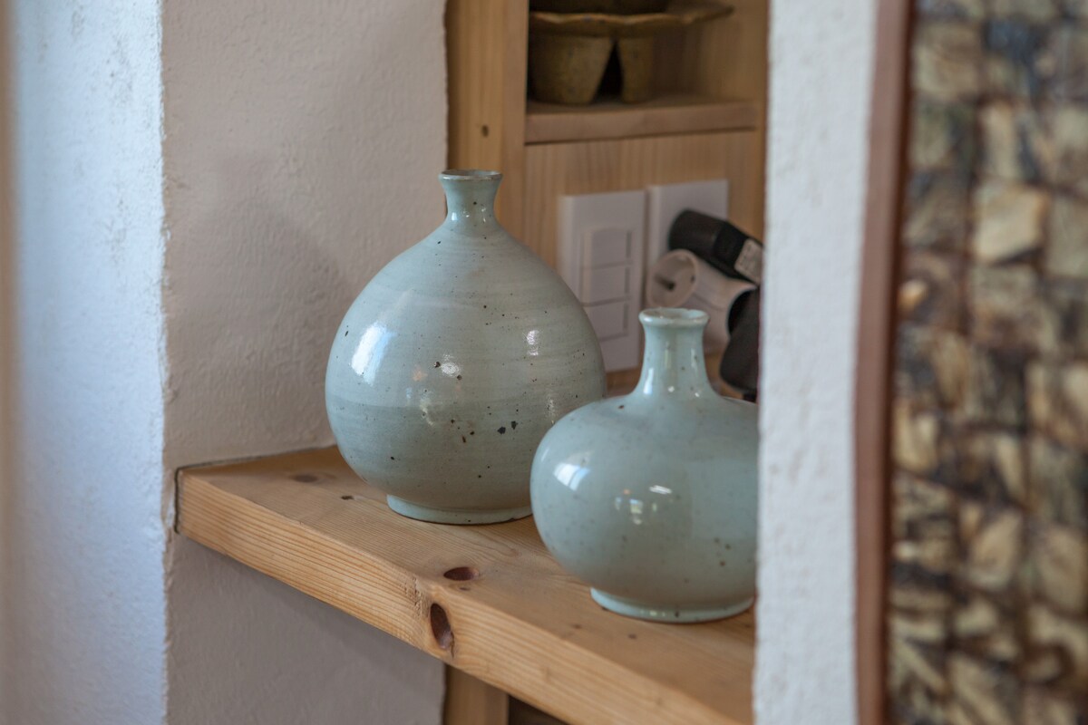 Two ceramic vases with smooth surfaces are placed on a wooden shelf. One vase is larger and rounder, while the other is smaller and more elongated. Both feature a light blue-green glaze that reflects subtle variations in shade.