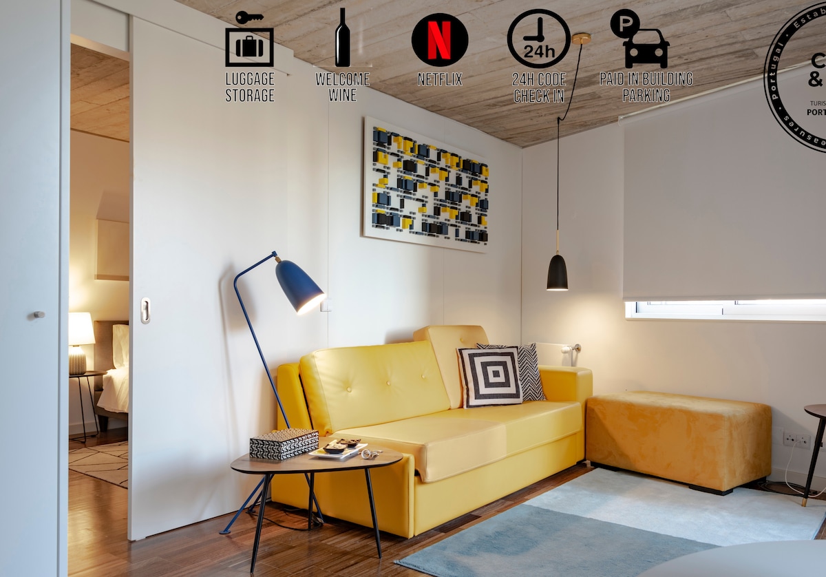 A modern living area is featured, showcasing a yellow sofa paired with a geometric-patterned throw pillow. A coffee table rests nearby, and a floor lamp provides soft lighting. The area is complemented by a vibrant piece of wall art and a large window allowing natural light to enter.