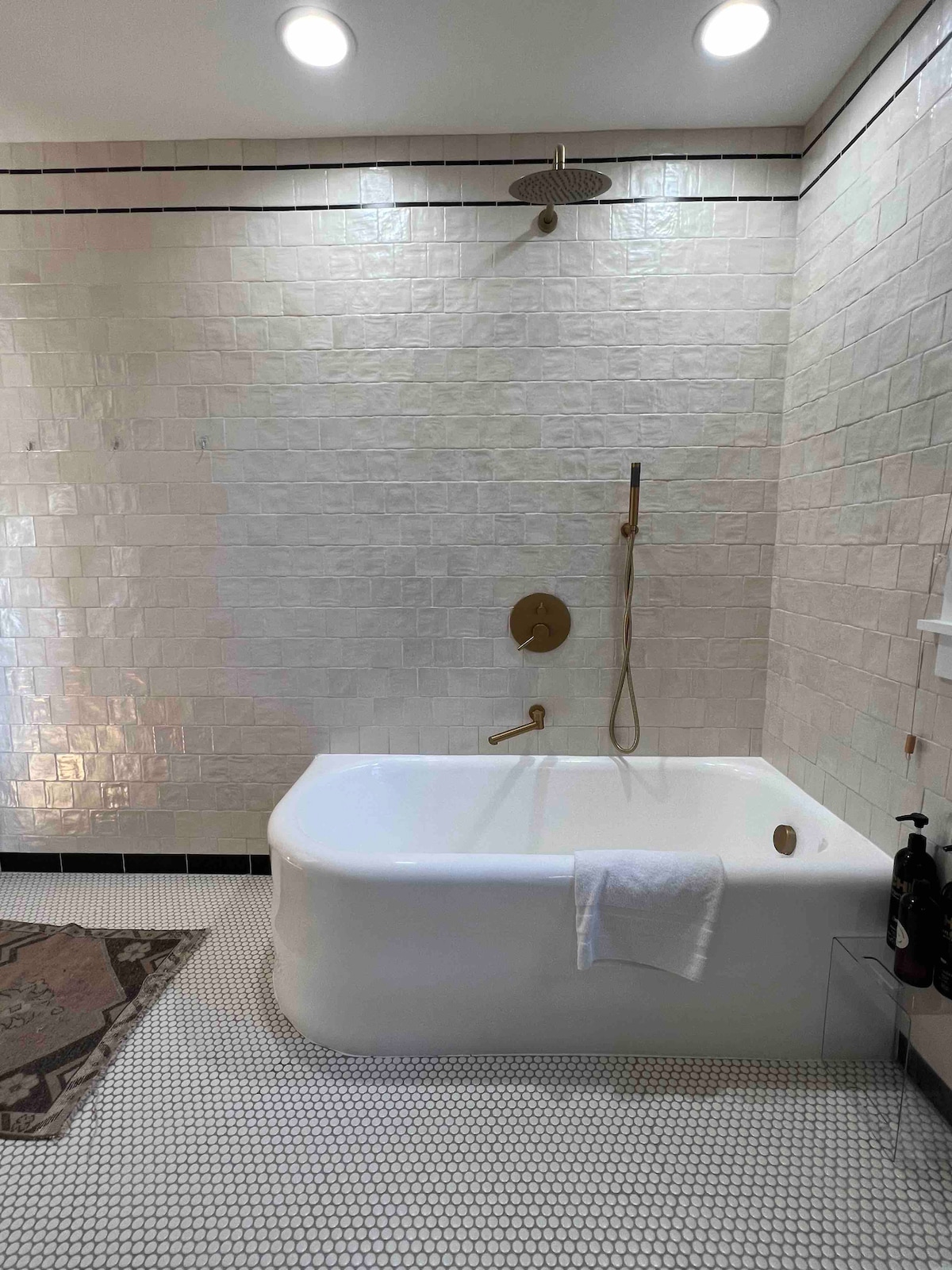 A spacious bathroom features a vintage cast-iron tub paired with a modern shower fixture. The walls are adorned with glossy tiles, and the floor is covered with distinctive mosaic patterns. Natural light illuminates the space from strategically placed ceiling lights.