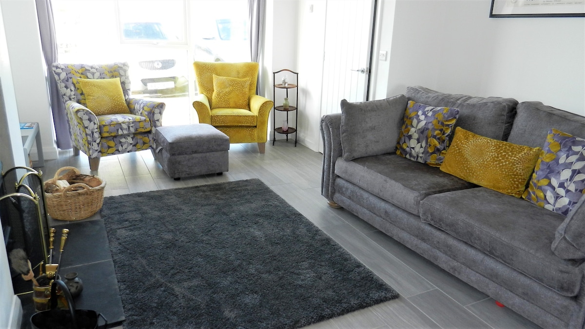 A modern living room features a grey sofa and two patterned armchairs with yellow accents. A grey area rug sits atop the tiled floor, while a small footstool complements the seating. A firewood holder and decorative items are positioned beside a wood burning stove.