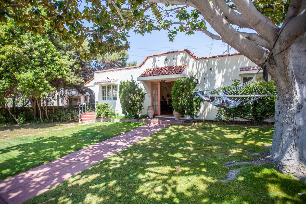 The front yard features lush green grass and a paved pathway leading to the entrance of a Spanish-style cottage. A hammock hangs casually between two trees, with well-maintained shrubs and foliage surrounding the area.
