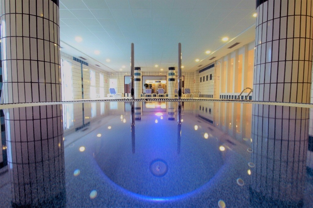 The indoor spa area features a heated pool with a reflective surface, surrounded by tiled columns. Soft lighting creates a serene ambiance, while lounge chairs are positioned nearby for relaxation. The overall design emphasizes a calming and inviting space for guests.