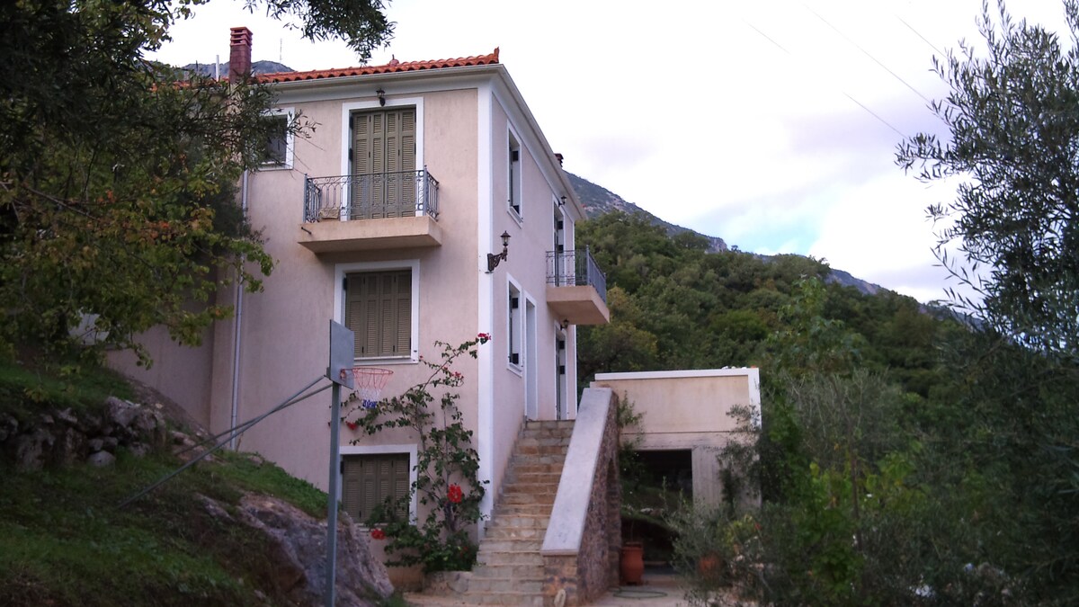 A two-story building is nestled among greenery, showcasing a light-colored exterior with a red-tiled roof. A stone staircase leads to the entrance, flanked by climbing greenery and flowering plants. The mountain backdrop adds to the natural setting of the home.
