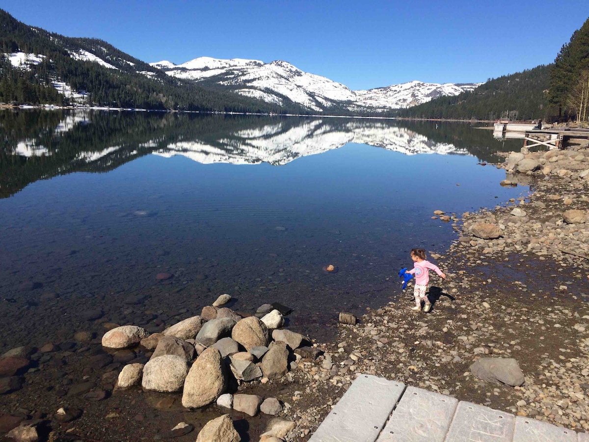 A child is seen playing near a calm lake, surrounded by a landscape of pine trees and mountains in the background. The clear water reflects the mountains and sky, while smooth stones line the shoreline.