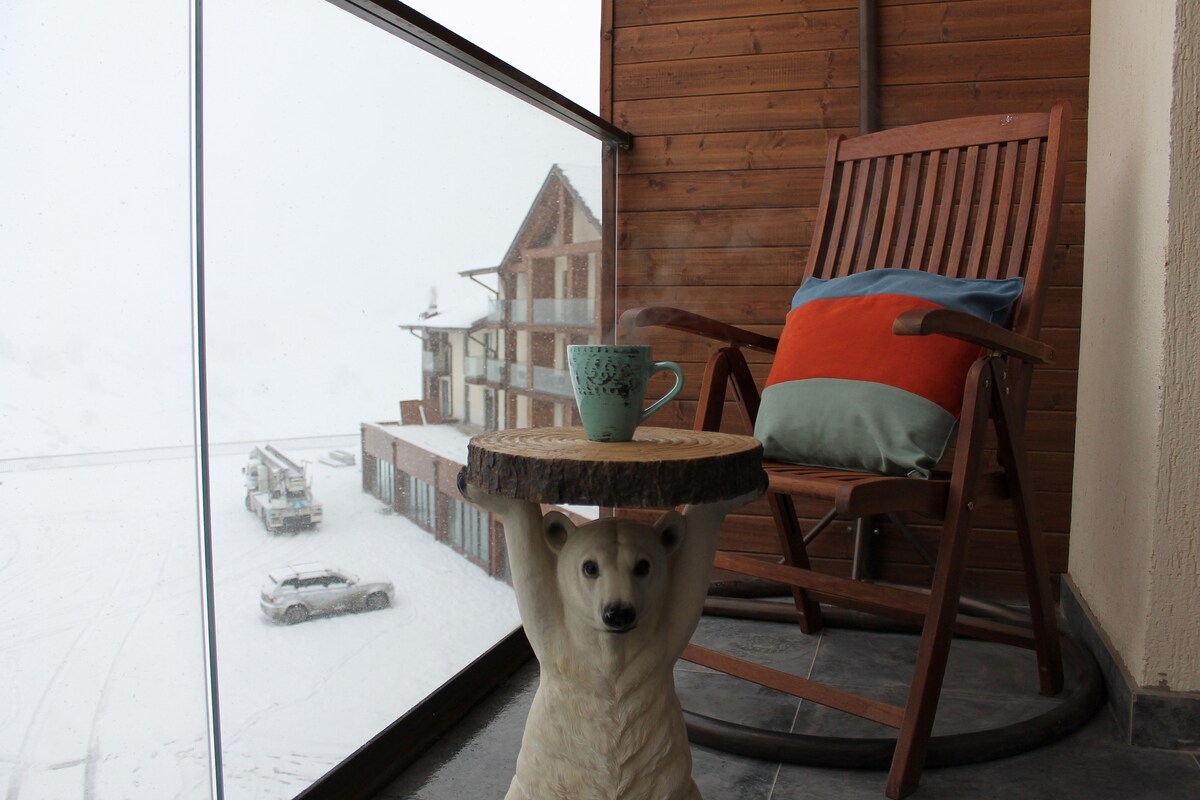 A cozy balcony is featured, featuring a wooden rocking chair with a colorful cushion next to a small table supported by a bear figurine. A cup rests on the table, with a snowy landscape visible in the background, highlighting the mountainous area.