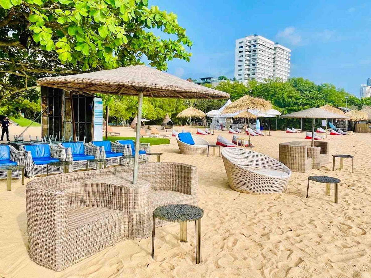 The beach area features various seating options arranged in a sandy environment. Woven lounge chairs and circular couches are shaded by umbrellas. The backdrop includes lush greenery and modern structures, while several cabanas are visible under the clear blue sky.