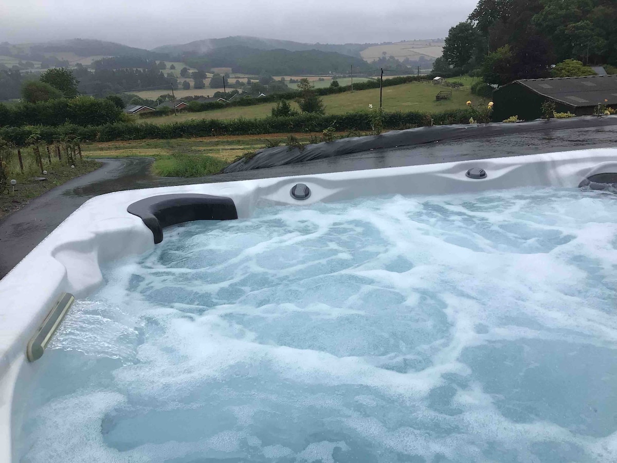 A hot tub filled with bubbling water is visible, set against a backdrop of rolling green hills and distant valleys. The sky appears cloudy, adding a serene ambiance to the scenic view.